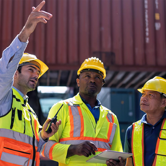 Three construction workers in safety vests and hard hats discussing a project at a construction site, with one worker pointing and another holding a tablet.