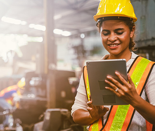 Smiling woman in a safety vest and hard hat using a tablet in an industrial setting.